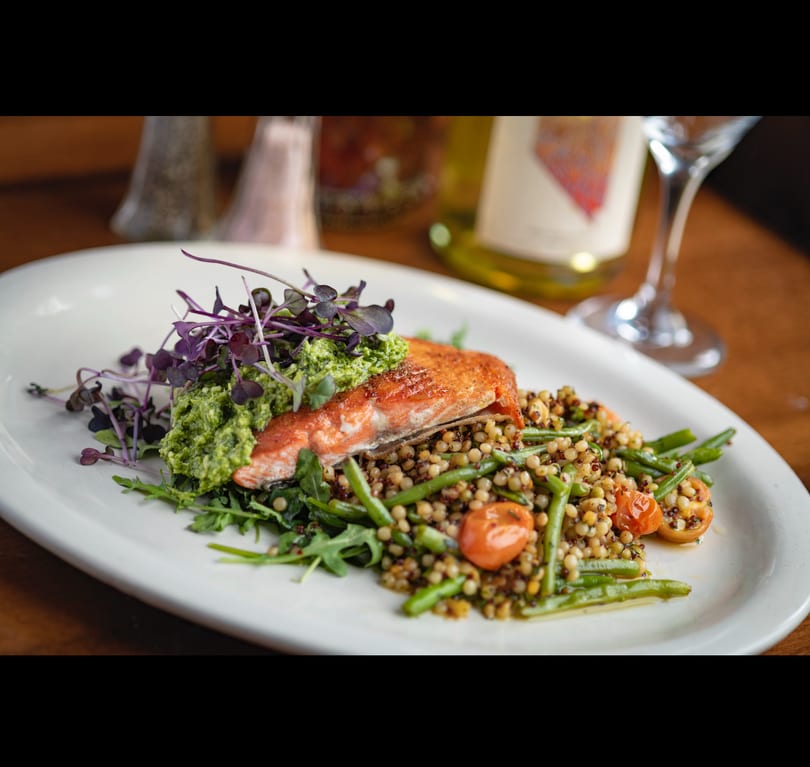 Pan-seared salmon topped with pesto and microgreens, served with pearl couscous, green beans, and tomatoes.