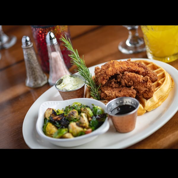 Fried chicken and waffles with roasted Brussels sprouts, sauces, and rosemary garnish on a white plate.