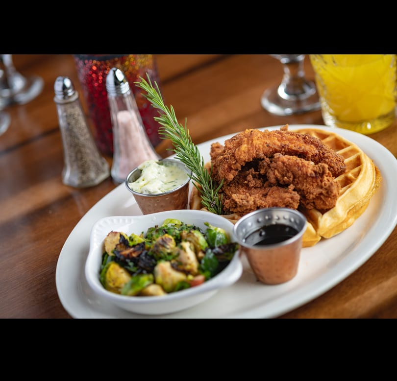 Fried chicken and waffles with roasted Brussels sprouts, sauces, and rosemary garnish on a white plate.