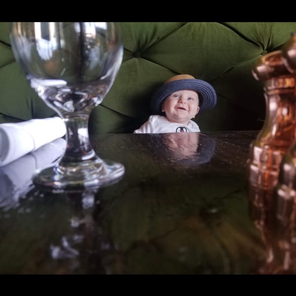 Happy baby in a straw hat sitting in a green restaurant booth with a wine glass.