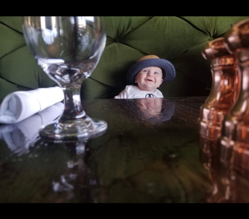 Happy baby in a straw hat sitting in a green restaurant booth with a wine glass.