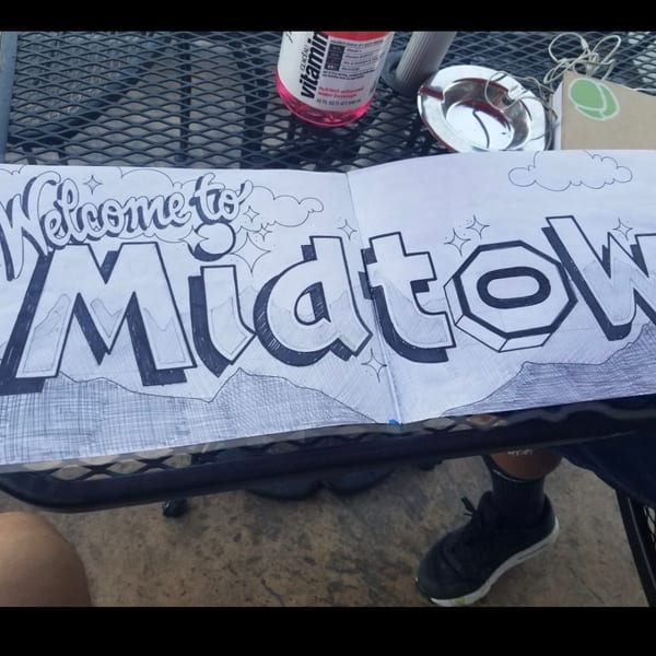 Stylized 'Welcome to Midtown' drawing on paper, with mountains and clouds, on an outdoor table.
