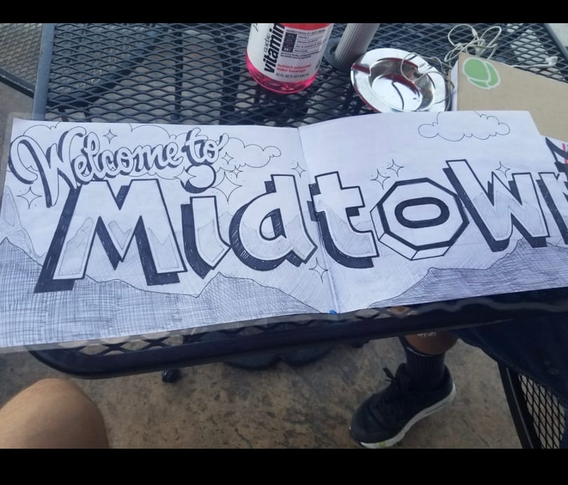 Stylized 'Welcome to Midtown' drawing on paper, with mountains and clouds, on an outdoor table.