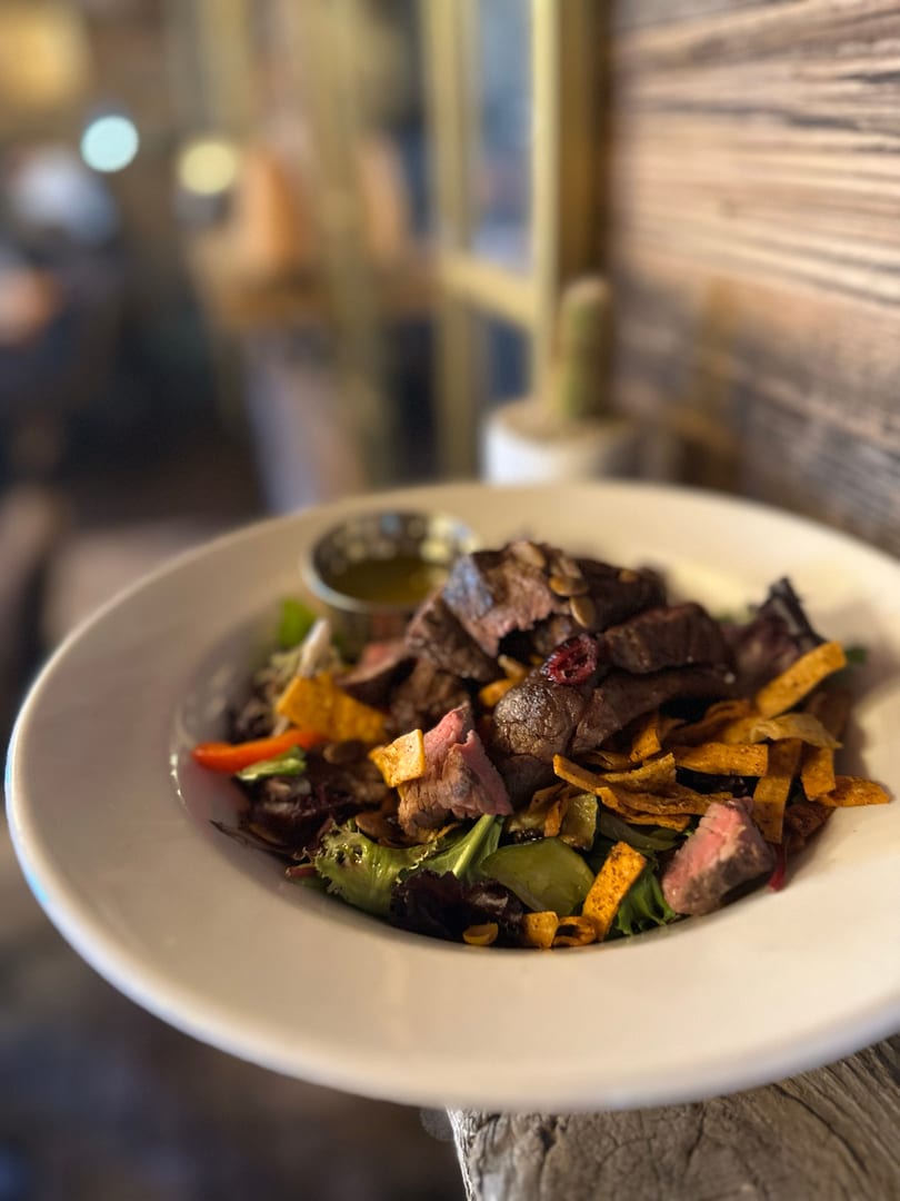 Colorful steak salad with mixed greens, tortilla strips, pumpkin seeds, and a side of dressing.