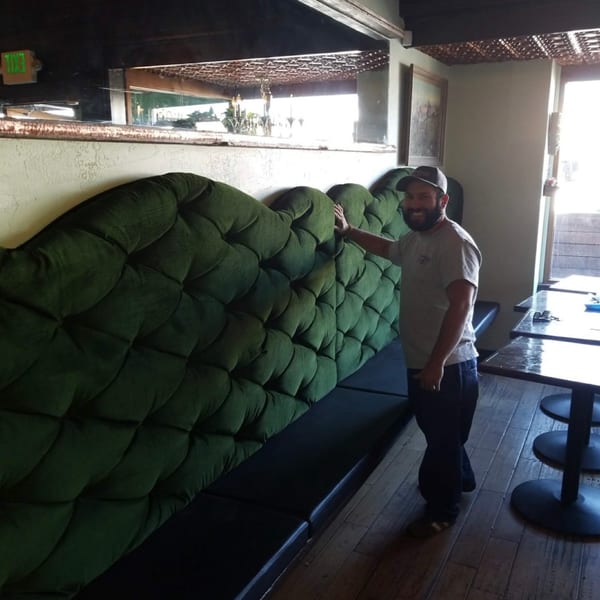 Man smiling next to a long, tufted green booth in a restaurant with wooden floors.