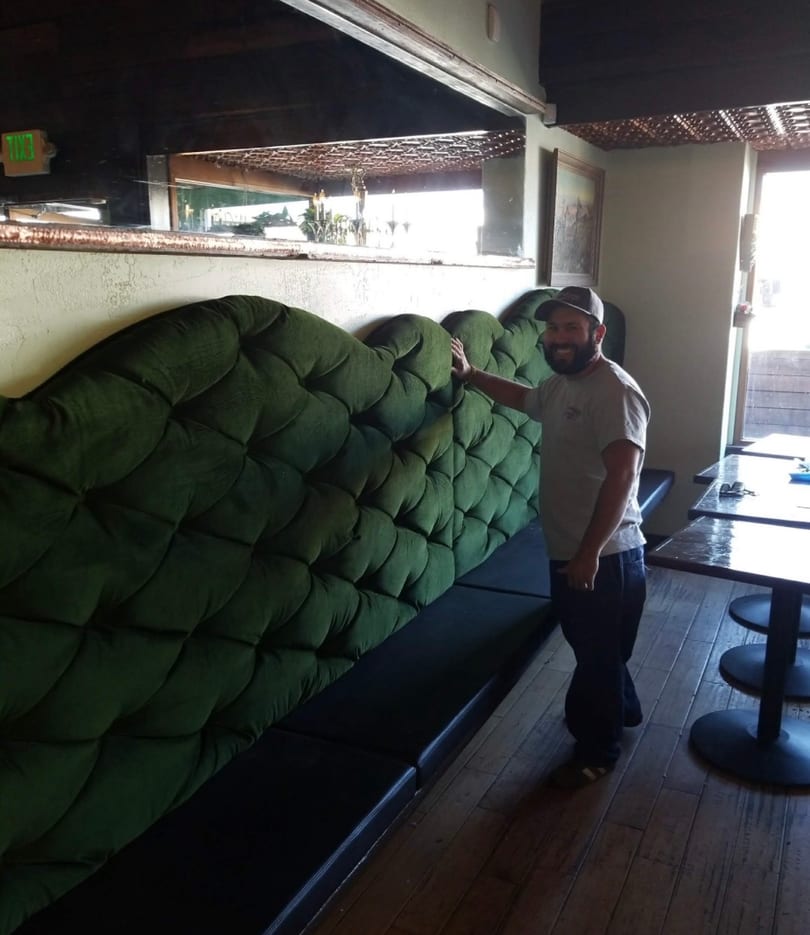 Man smiling next to a long, tufted green booth in a restaurant with wooden floors.