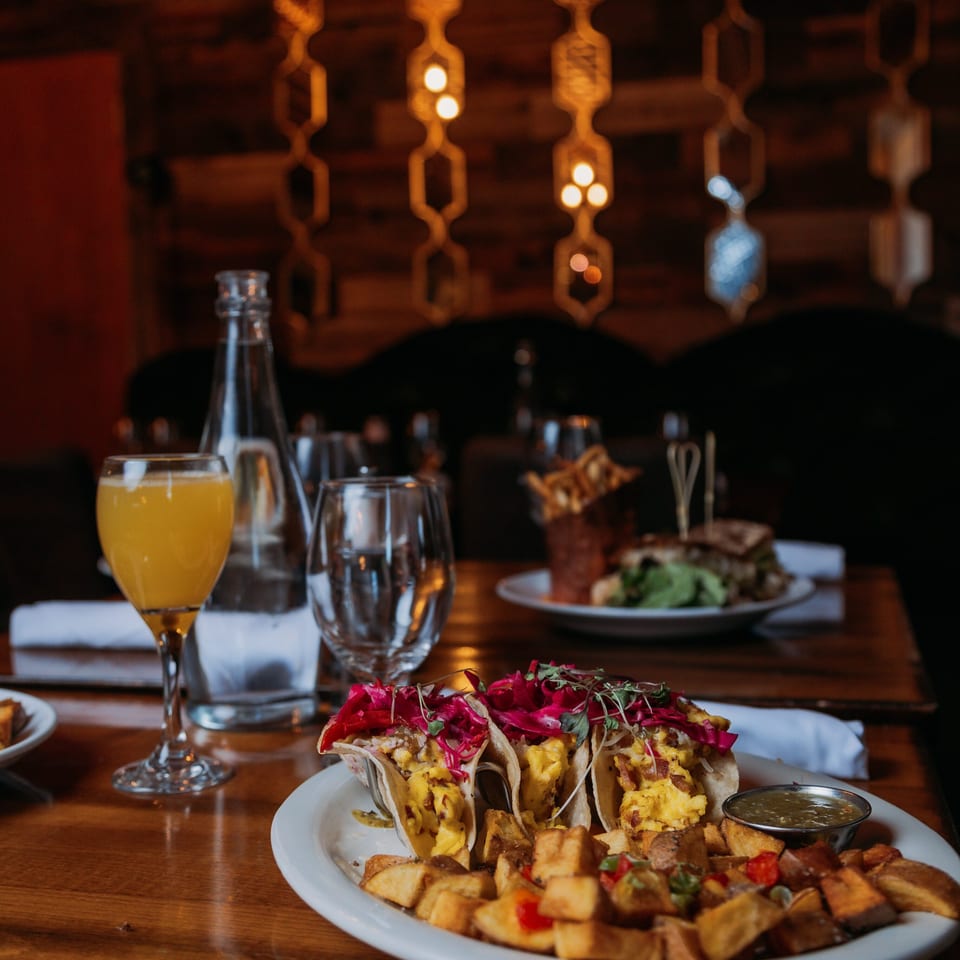 Scrambled egg tacos with red cabbage, home fries, and a mimosa on a restaurant table.