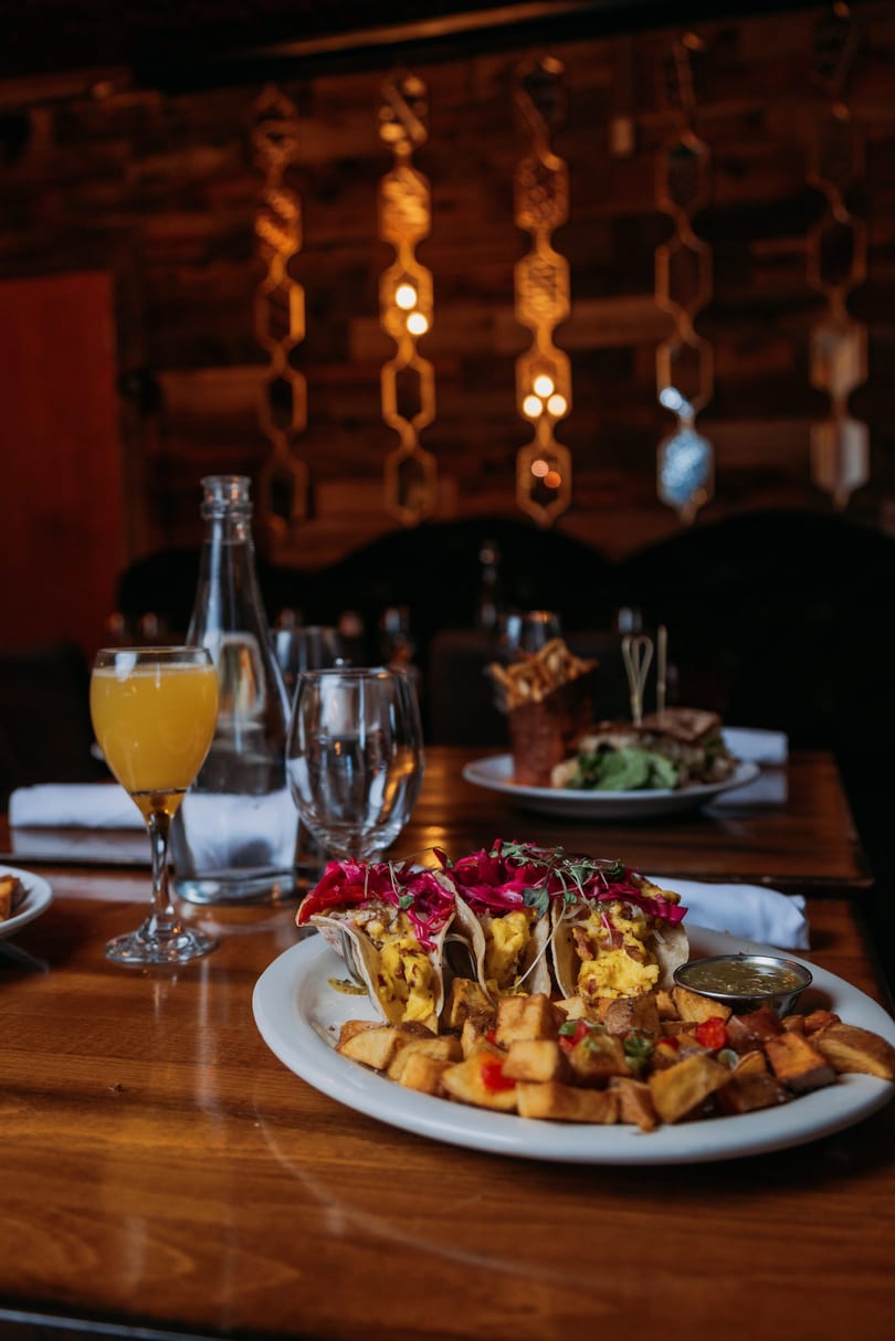 Scrambled egg tacos with red cabbage, home fries, and a mimosa on a restaurant table.