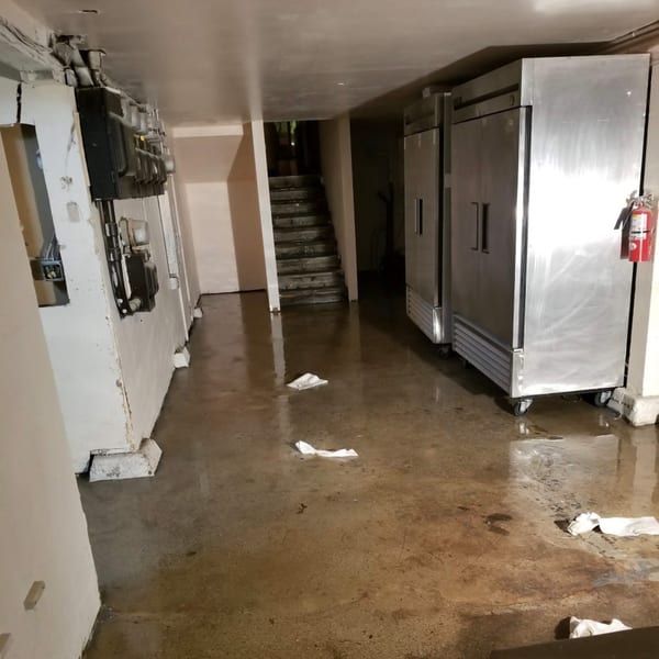 Flooded restaurant basement with stainless steel refrigerators, electrical meters, and stairs.