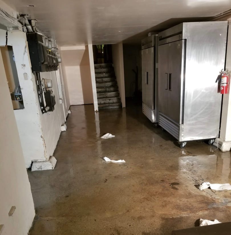 Flooded restaurant basement with stainless steel refrigerators, electrical meters, and stairs.