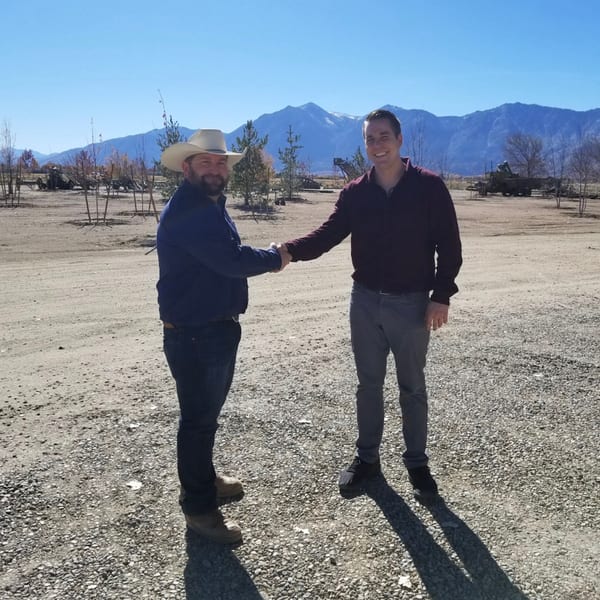 Two men shaking hands on a gravel road with mountains and trees under a clear sky.