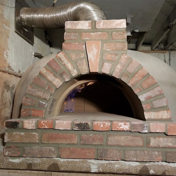 Arched brick and concrete pizza oven with a blue flame inside and a metal exhaust vent.