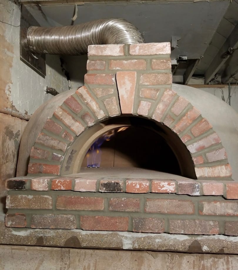 Arched brick and concrete pizza oven with a blue flame inside and a metal exhaust vent.