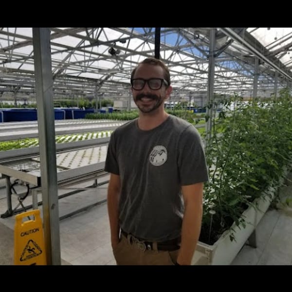 Smiling man in glasses and a mustache standing in a modern hydroponic greenhouse.