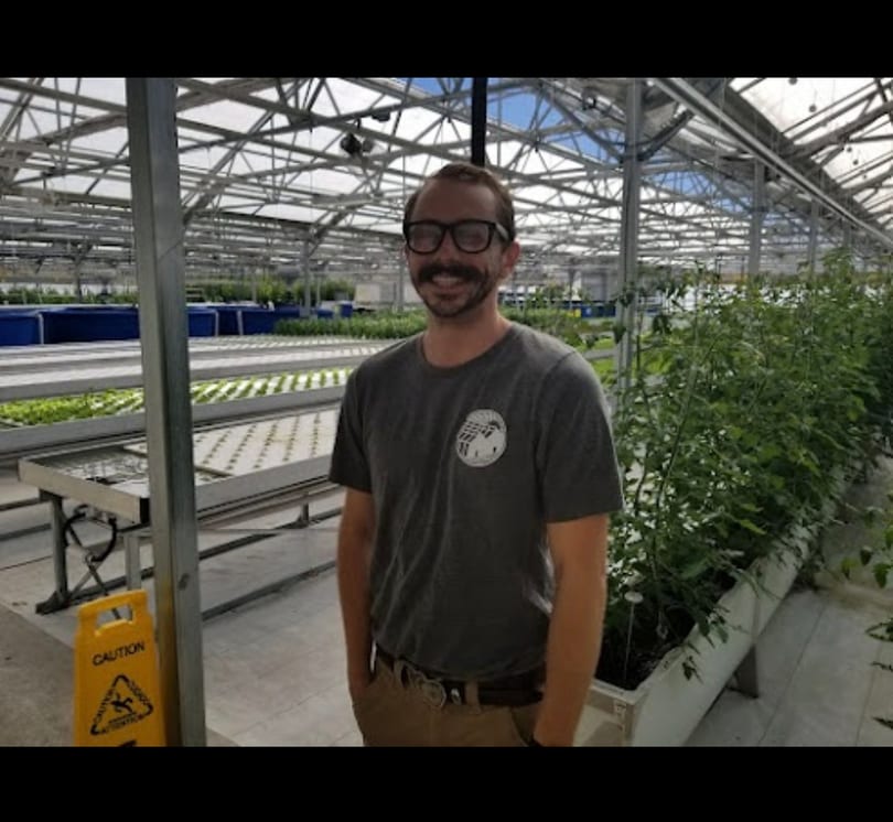 Smiling man in glasses and a mustache standing in a modern hydroponic greenhouse.
