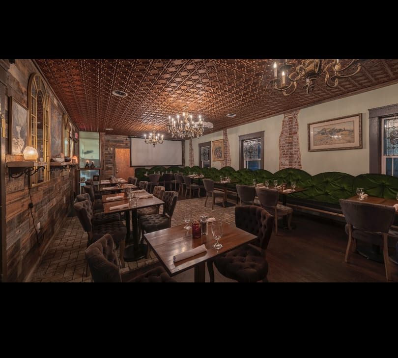 Elegant dining room with plush chairs, green booths, ornate chandeliers, and a projector screen.