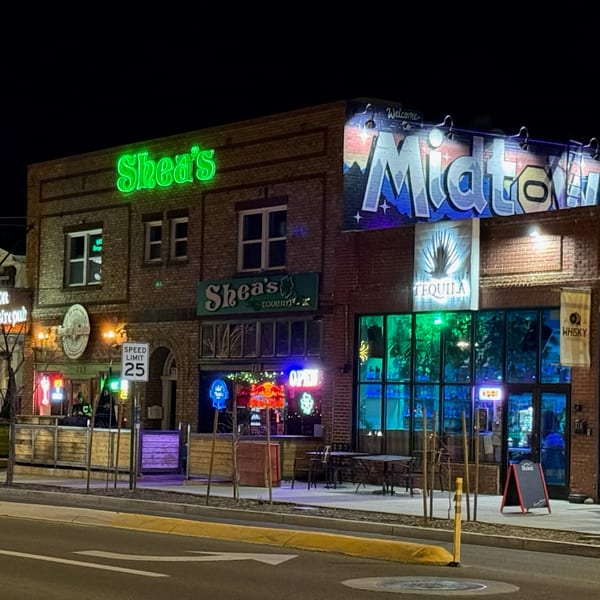 Night view of brick buildings with neon signs for Shea's, Homegrown Gastropub, and Midtown, featuring outdoor seating.