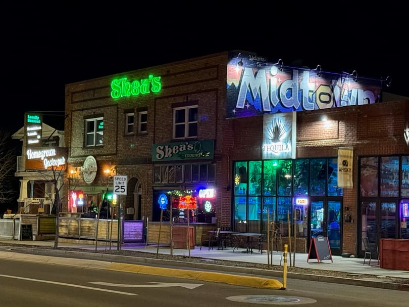 Night view of brick buildings with neon signs for Shea's, Homegrown Gastropub, and Midtown, featuring outdoor seating.
