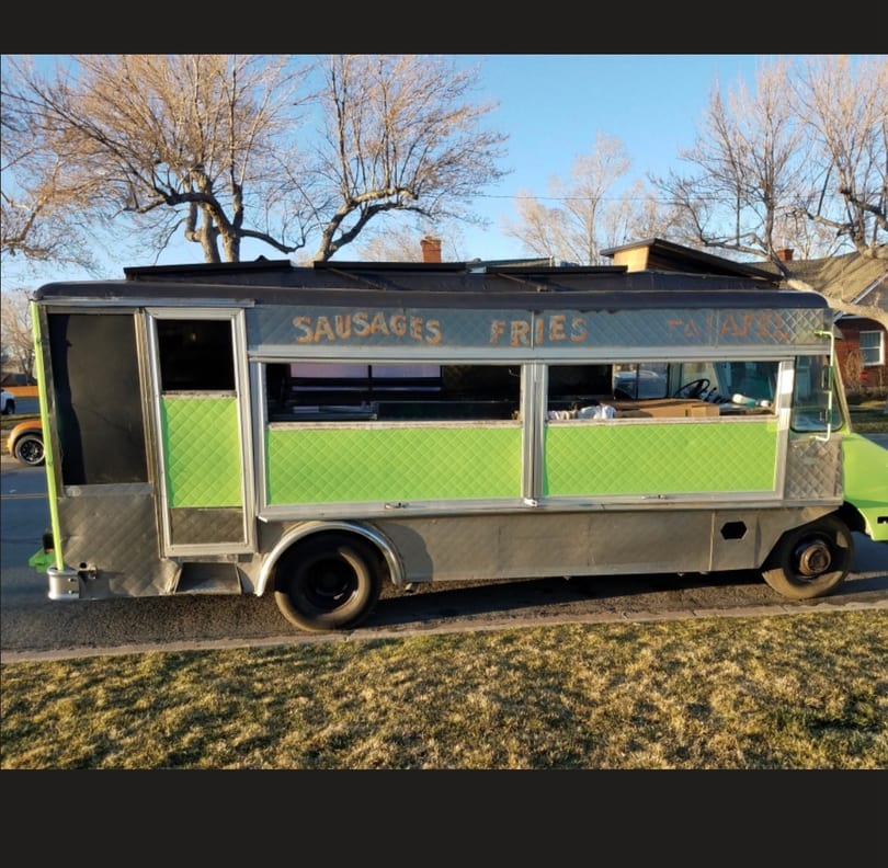 Gray and lime green food truck with 'SAUSAGES FRIES' on the side, parked on grass.