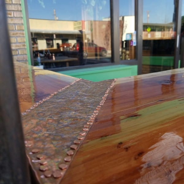 Shiny wooden bar top with hammered copper inlay and rivets, reflecting an outdoor street scene.