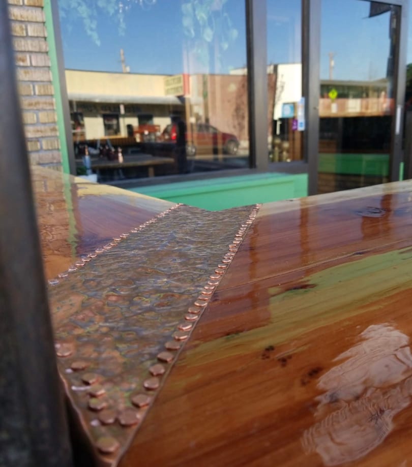 Shiny wooden bar top with hammered copper inlay and rivets, reflecting an outdoor street scene.