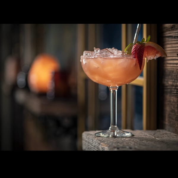A pink cocktail with a salted rim, ice, strawberry, lime, and grapefruit on a rustic wooden surface.