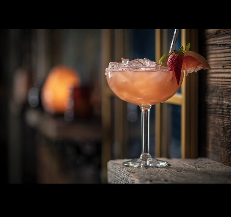 A pink cocktail with a salted rim, ice, strawberry, lime, and grapefruit on a rustic wooden surface.