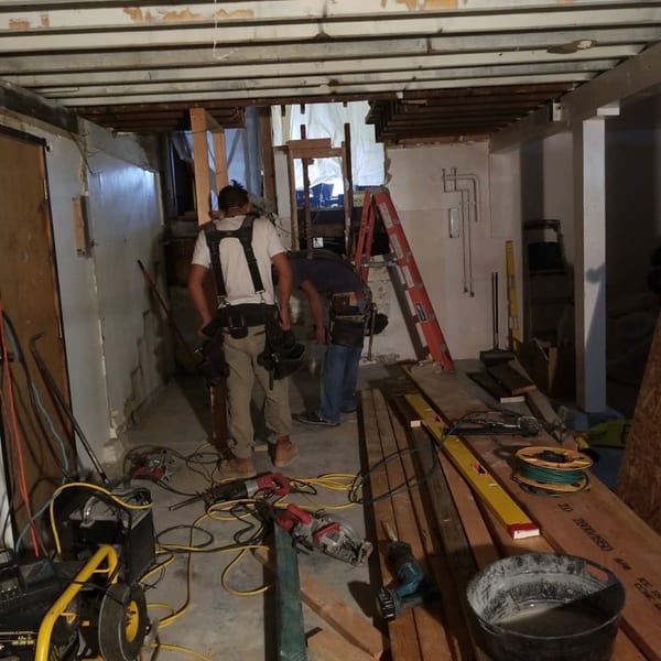Two construction workers in a room under renovation with tools and exposed ceiling joists.