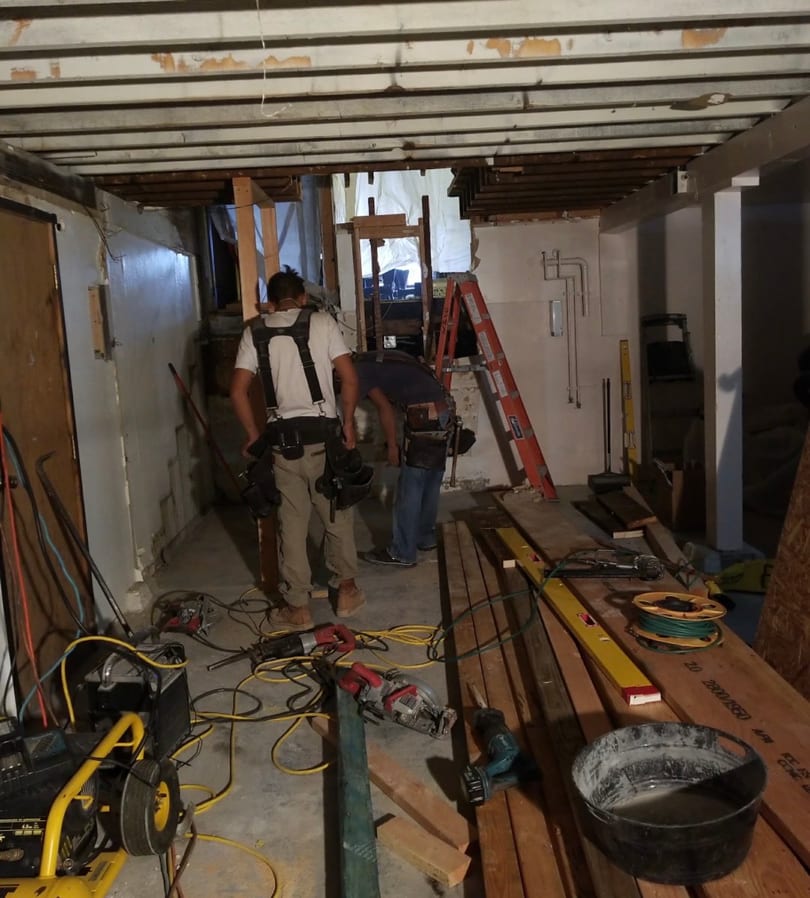 Two construction workers in a room under renovation with tools and exposed ceiling joists.