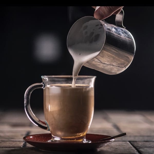 Hand pouring frothed milk from a metal pitcher into a glass mug of coffee on a wooden table.