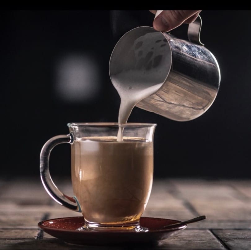 Hand pouring frothed milk from a metal pitcher into a glass mug of coffee on a wooden table.