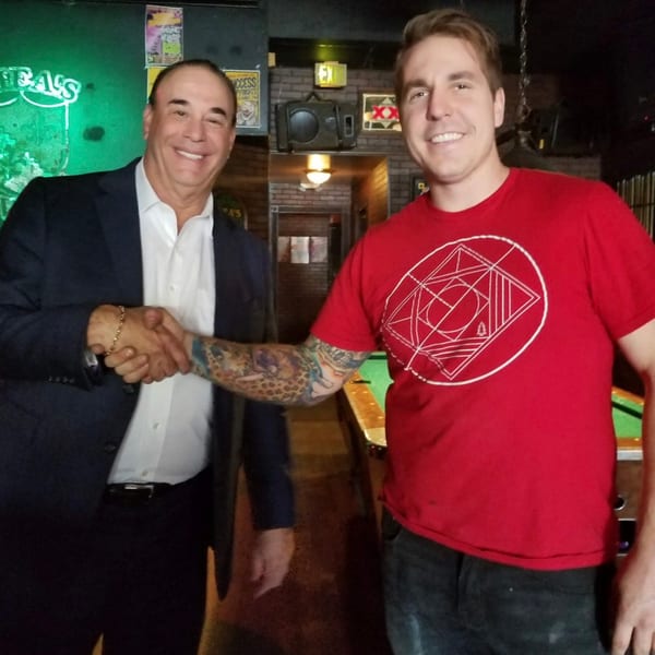 Two smiling men shake hands in a bar, with a pool table visible.