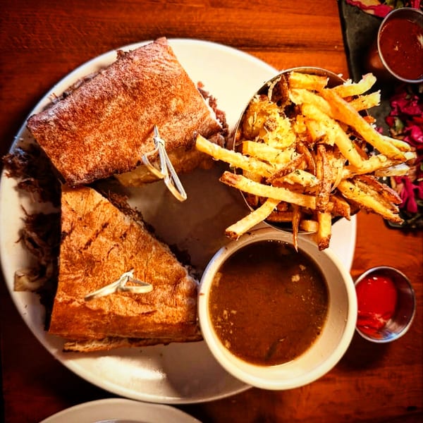 French dip sandwich with crispy fries, au jus, and ketchup on a wooden table.
