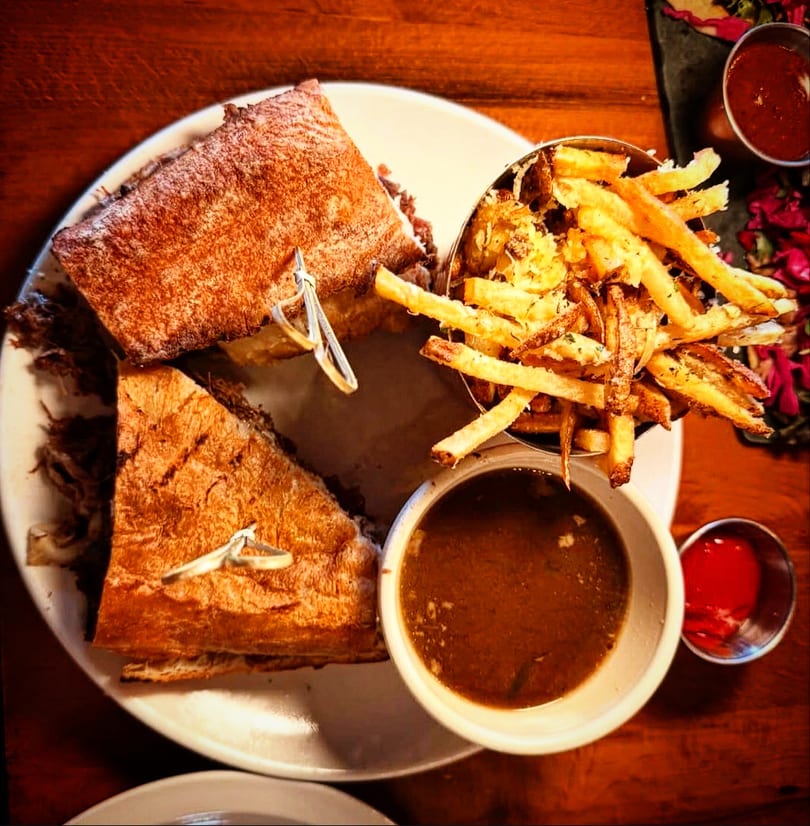 French dip sandwich with crispy fries, au jus, and ketchup on a wooden table.