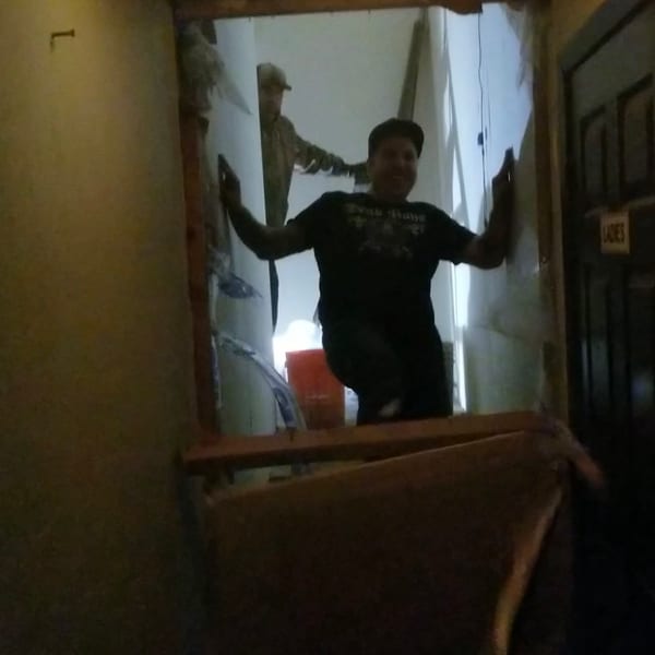 Two men in a dimly lit, unfinished doorway, one smiling, with a "LADIES" sign.