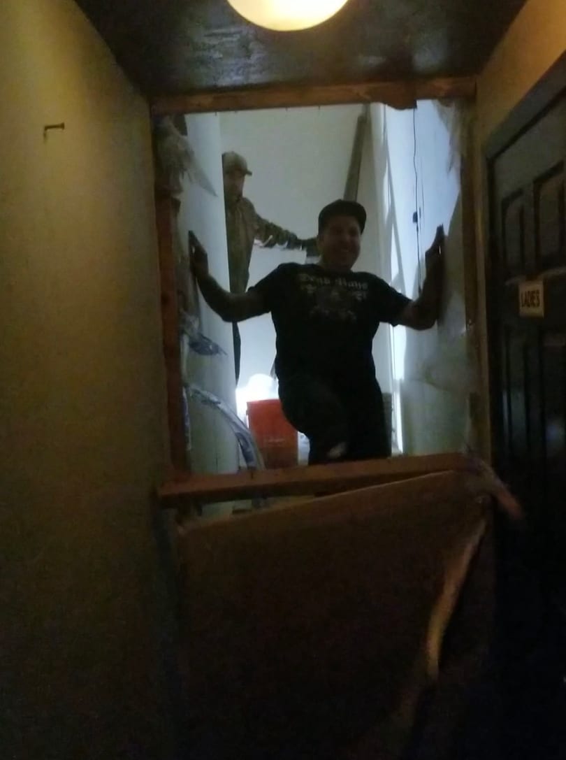 Two men in a dimly lit, unfinished doorway, one smiling, with a "LADIES" sign.