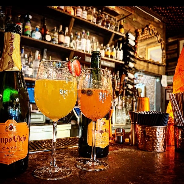 Two mimosa-style drinks and Campo Viejo Cava bottles on a rustic bar with liquor shelves.