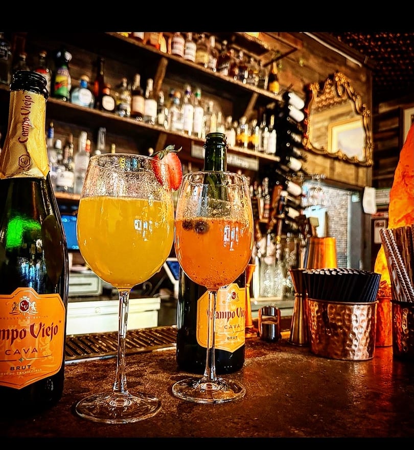 Two mimosa-style drinks and Campo Viejo Cava bottles on a rustic bar with liquor shelves.