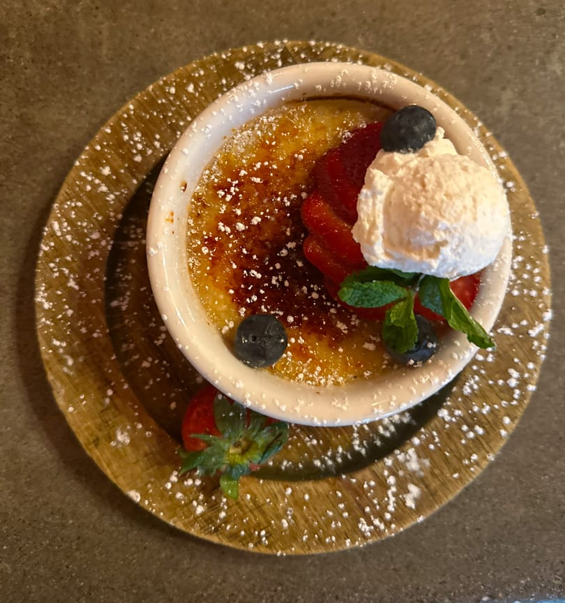 Caramelized creme brulee with whipped cream, strawberries, blueberries, and mint on a wooden plate.