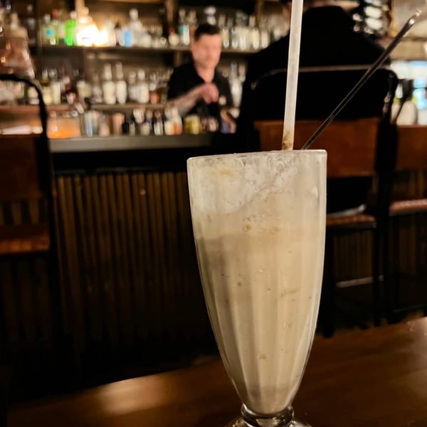Creamy drink with a straw on a dark bar table, with a bartender and bottles in the background.