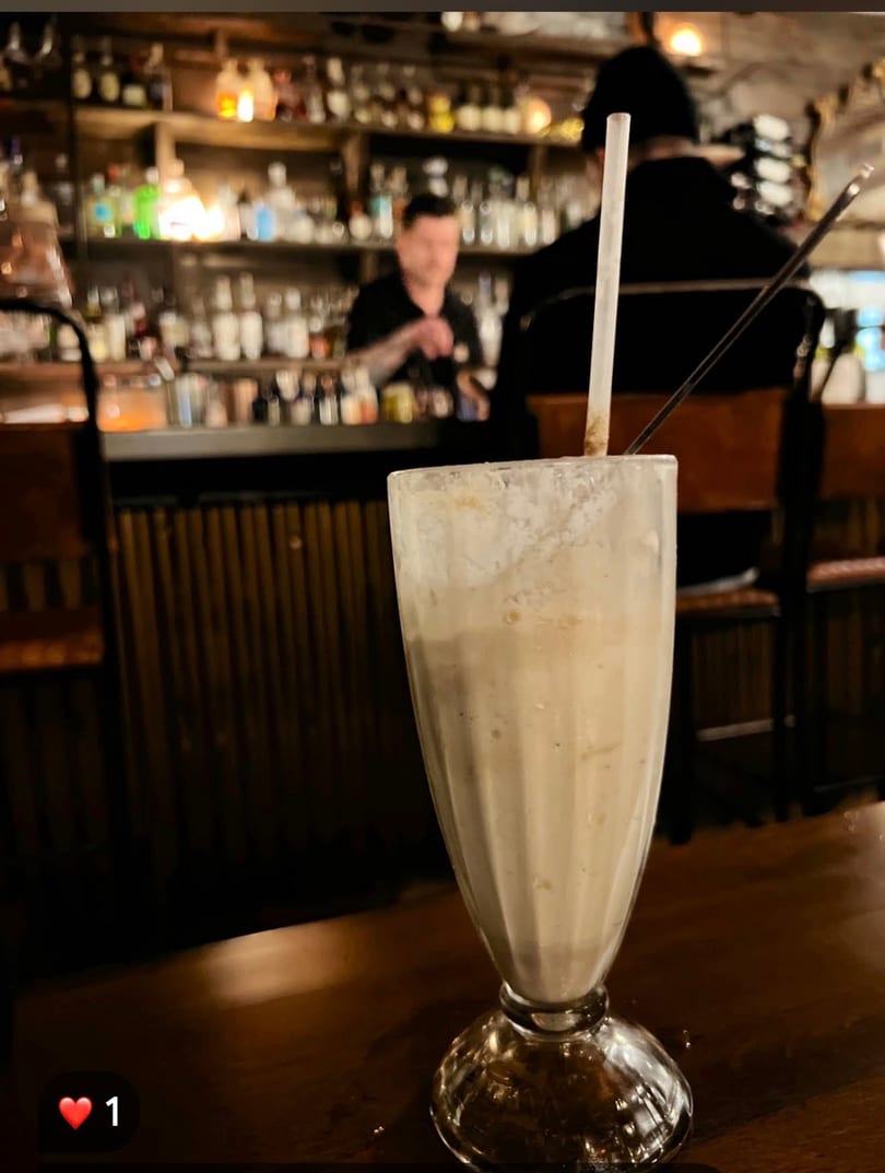 Creamy drink with a straw on a dark bar table, with a bartender and bottles in the background.