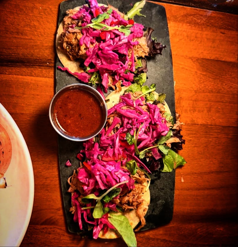 Overhead view of three tacos with pulled pork, vibrant purple cabbage, and dipping sauce.