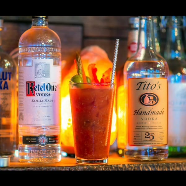 Garnished Bloody Mary cocktail with vodka bottles (Ketel One, Tito's) and warm backlighting.