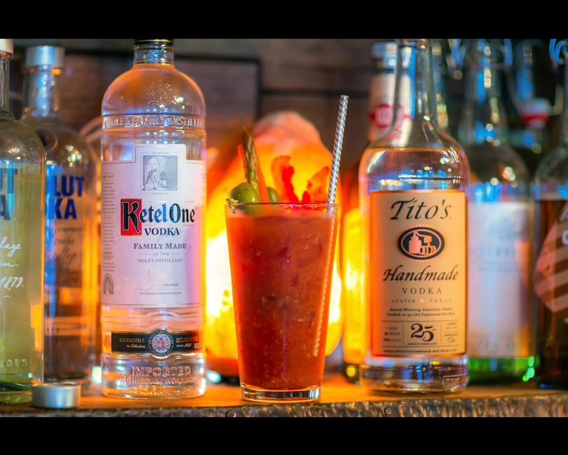 Garnished Bloody Mary cocktail with vodka bottles (Ketel One, Tito's) and warm backlighting.