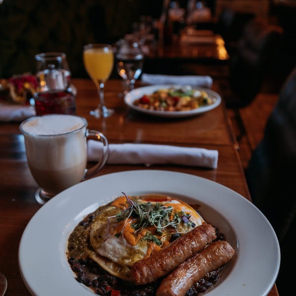 Breakfast plate with fried eggs, sausages, black beans, a latte, and orange juice on a wooden table.