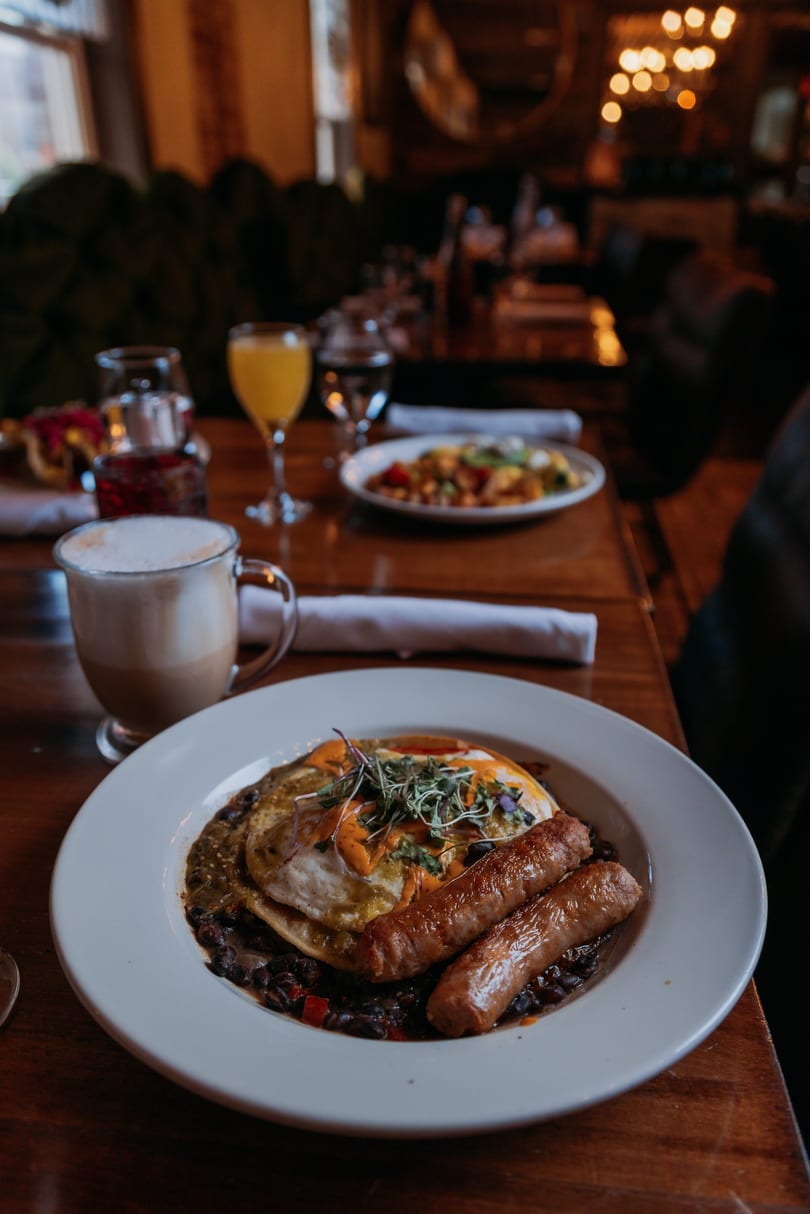 Breakfast plate with fried eggs, sausages, black beans, a latte, and orange juice on a wooden table.