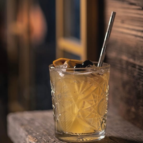A patterned glass cocktail with orange, cherries, and a metal straw on a rustic wooden surface.
