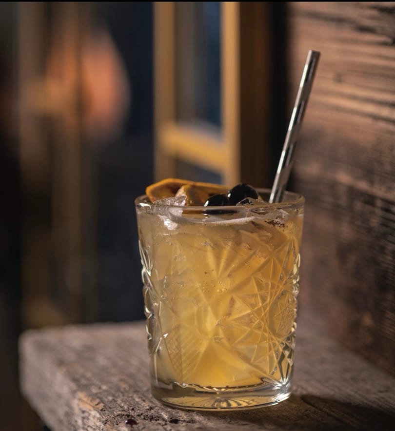 A patterned glass cocktail with orange, cherries, and a metal straw on a rustic wooden surface.