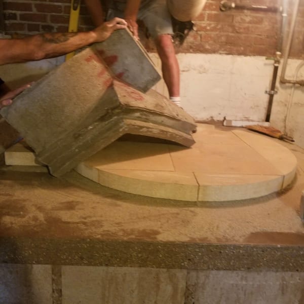 Two people construct a circular oven base with large stone pieces against a brick wall.