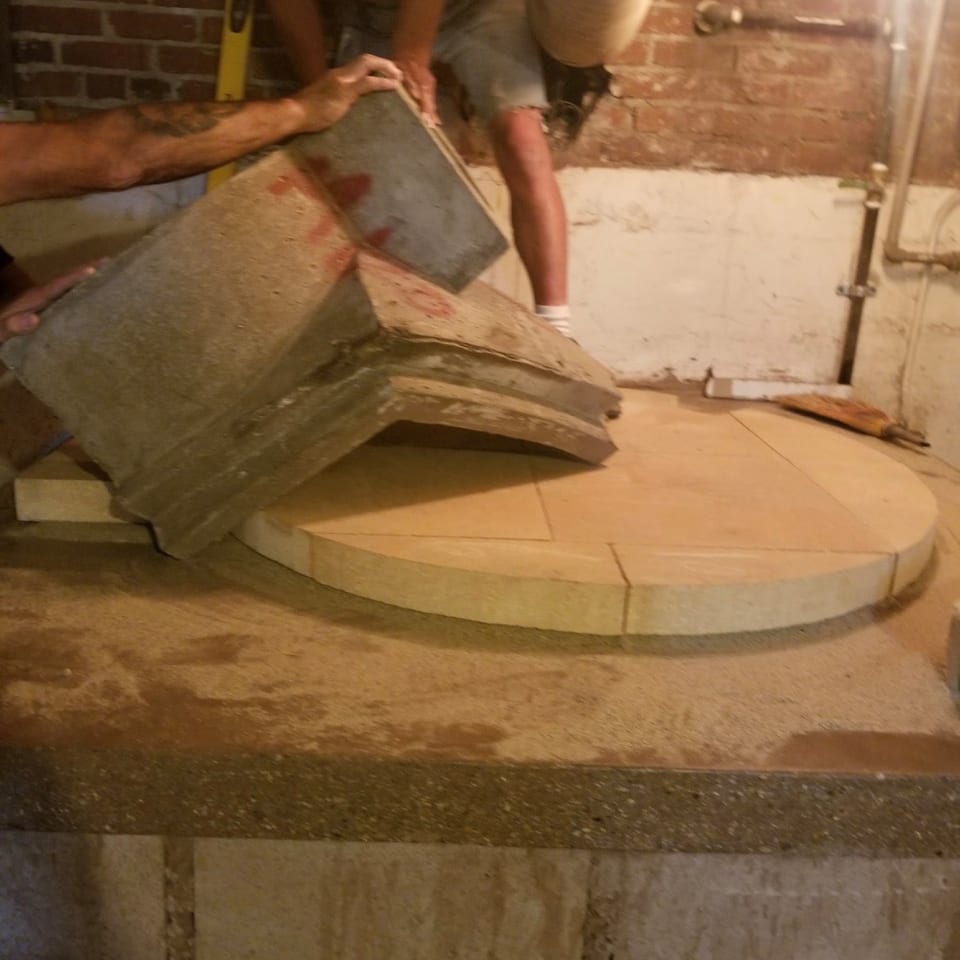 Two people construct a circular oven base with large stone pieces against a brick wall.